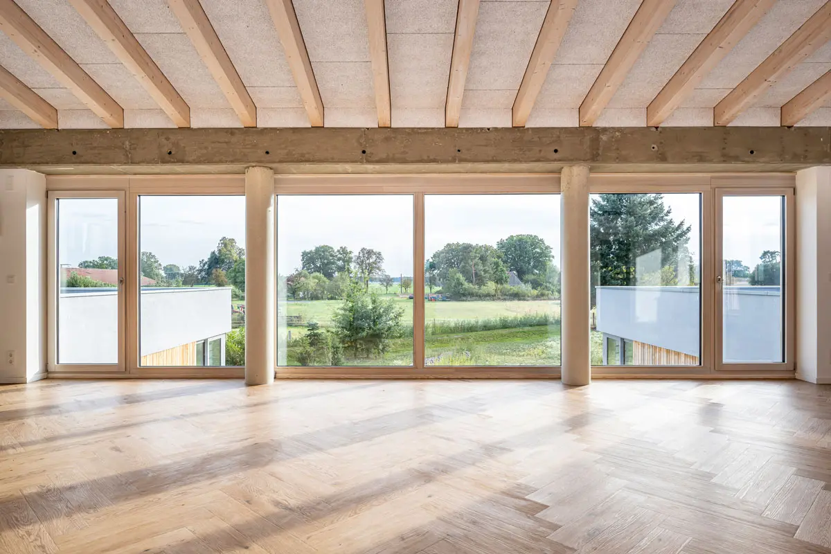 Heller Raum mit Parkett, Holzbalken und großer Fensterfront mit Blick auf Wiesen und Bäume