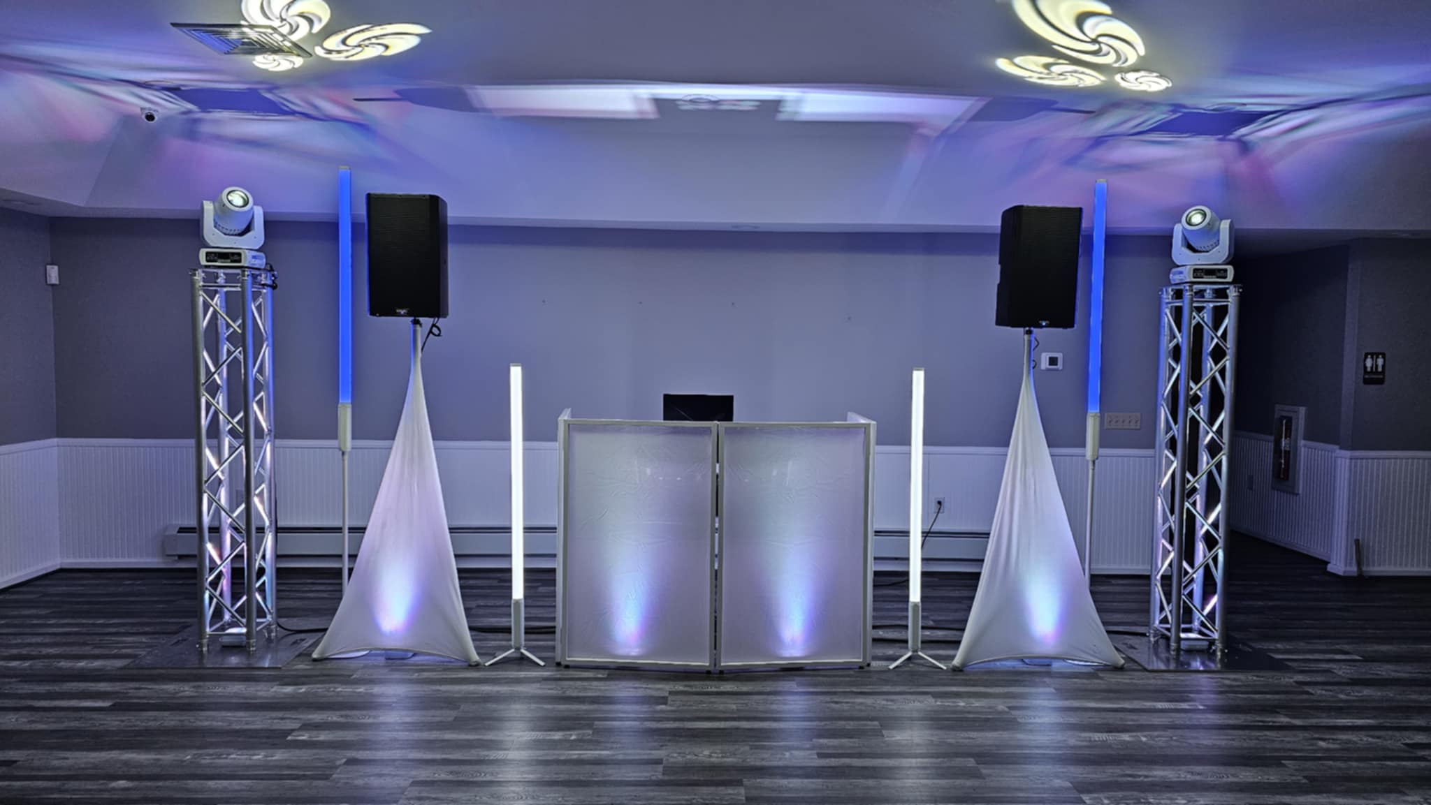 Two moving head lights on vertical truss throwing patterns across the ceiling behind a white DJ booth