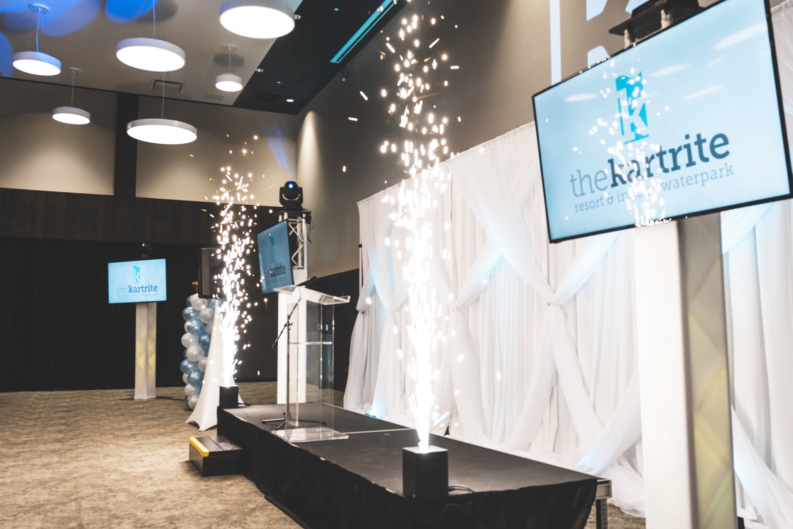 Corporate event stage with cold spark fountains, a podium, branded screens and white pipe and drape