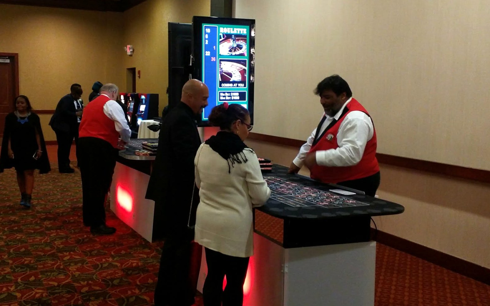 Roulette dealer with guests at an uplit roulette table beside a 32-inch screen tracking the last numbers spun at a corporate gala