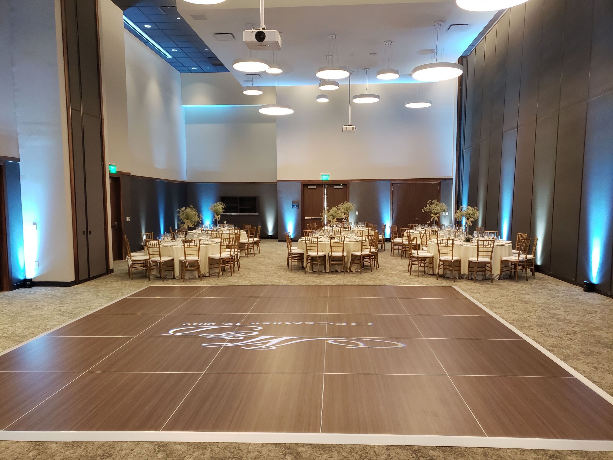 Ballroom walls washed in teal uplighting around dinner tables and a dance floor with a projected monogram