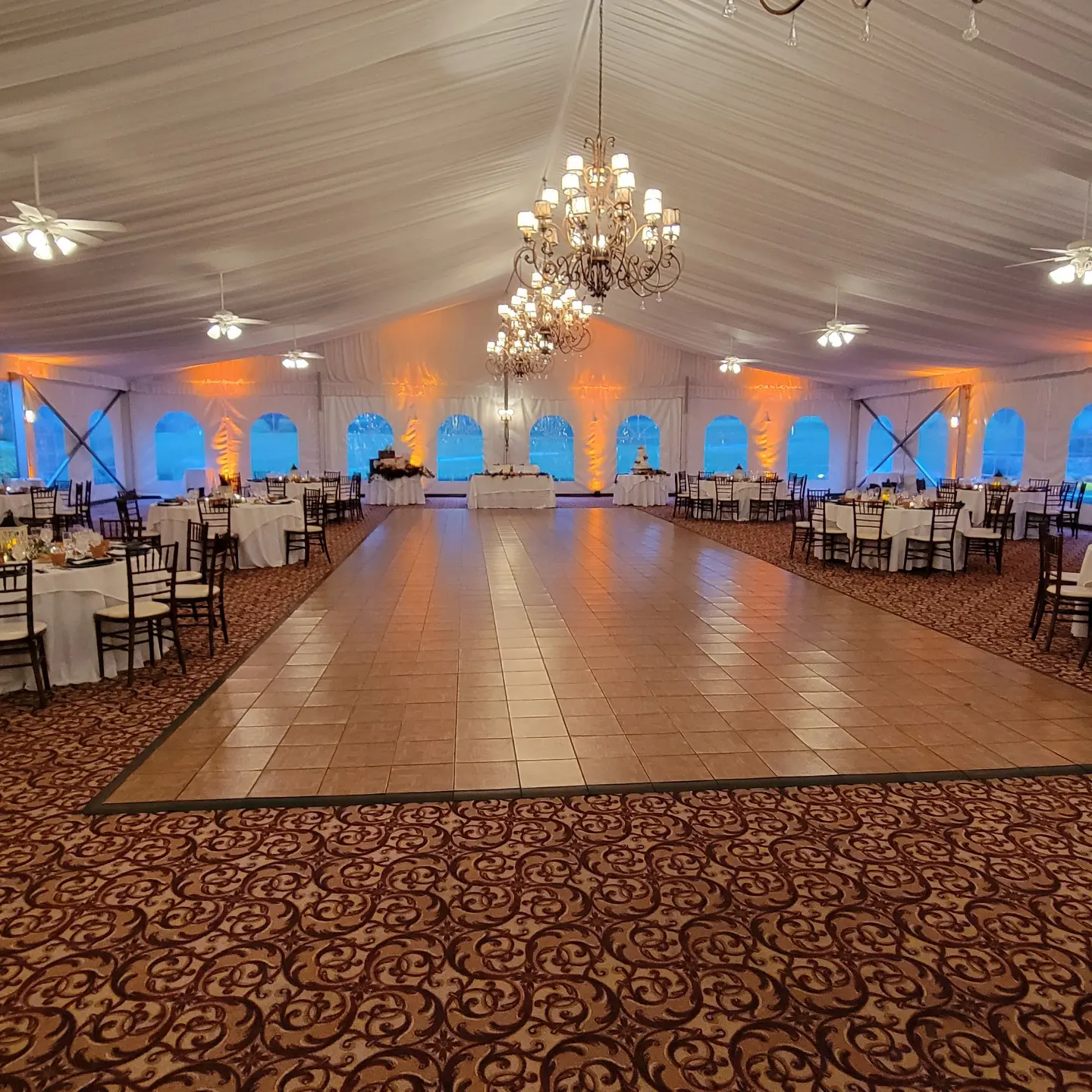 Wood dance floor in the middle of a white tent reception with amber uplighting and chandeliers