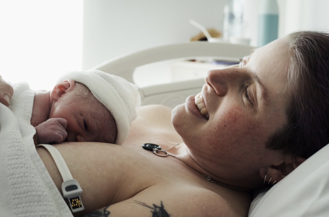A person with purple hair smiling in a hospital bed with a tiny baby on their chest. A person with purple hair smiling in a hospital bed with a tiny baby on their chest.
