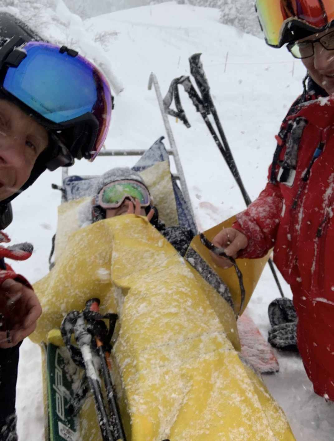 A barely visible person on a yellow sled with two mountain paramedics dressed in red. It's snowing. A barely visible person on a yellow sled with two mountain paramedics dressed in red. It's snowing.