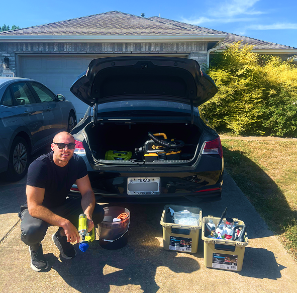 Car detailing technician kneeling beside a trunk filled with tools and cleaning supplies in a residential driveway in Texas.