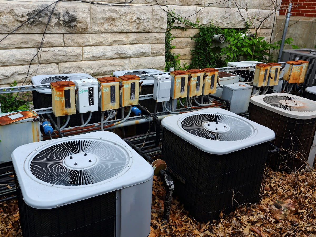 A series of aged outdoor air conditioning units with rusted electrical boxes, set against a stone wall, surrounded by overgrown plants.