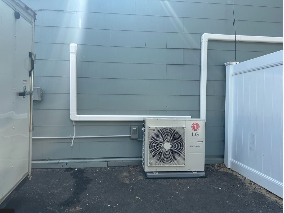 Outdoor LG Smart Inverter air conditioning unit installed against a house with gray siding, with pipes running along the wall.