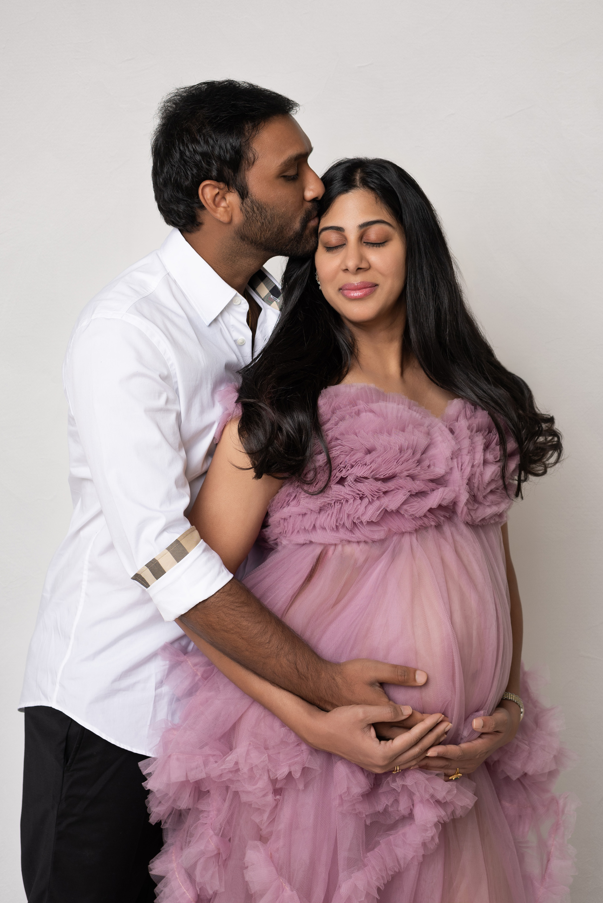 A pregnant woman in a purple gown being embraced by her husband