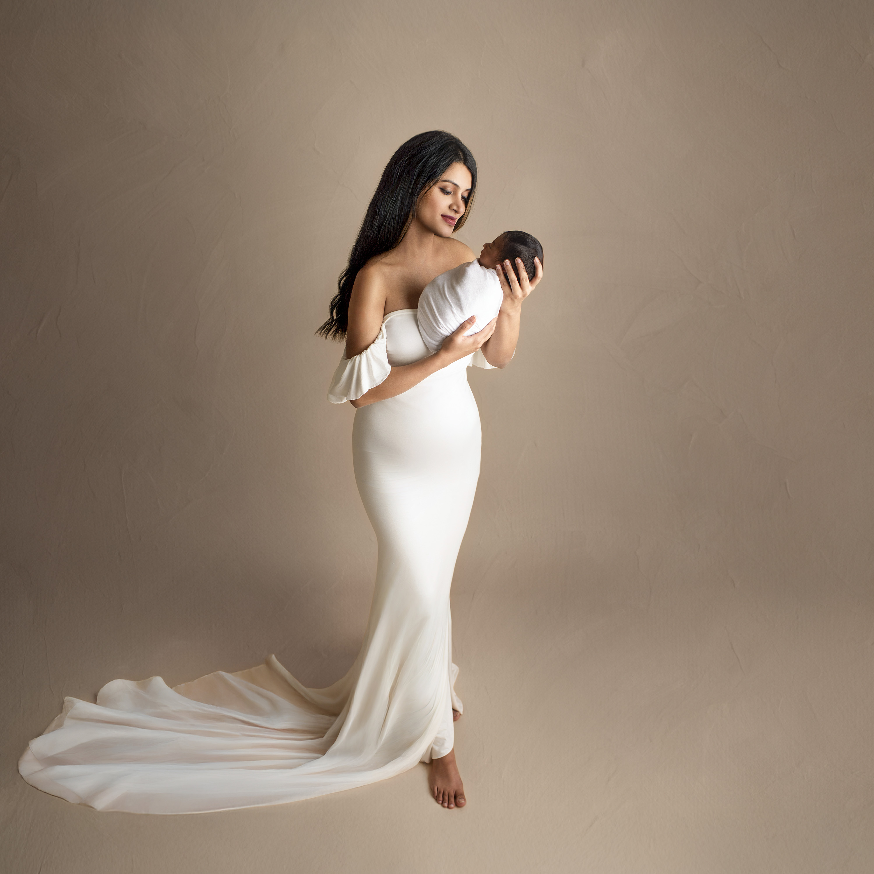 Mom wearing elegant white gown and holding a newborn baby boy swaddled in white. 