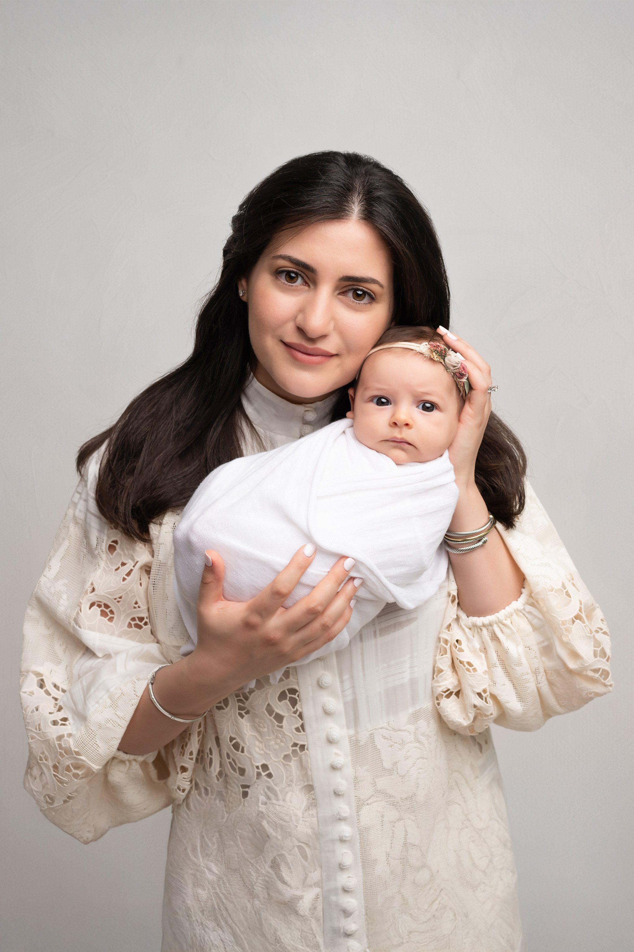 Mother holding newborn baby girl wrapped in white swaddle and smiling at the camera.