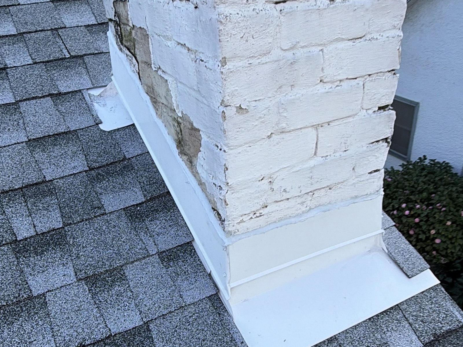 White metal chimney flashing at the base of a weathered brick chimney on gray shingles