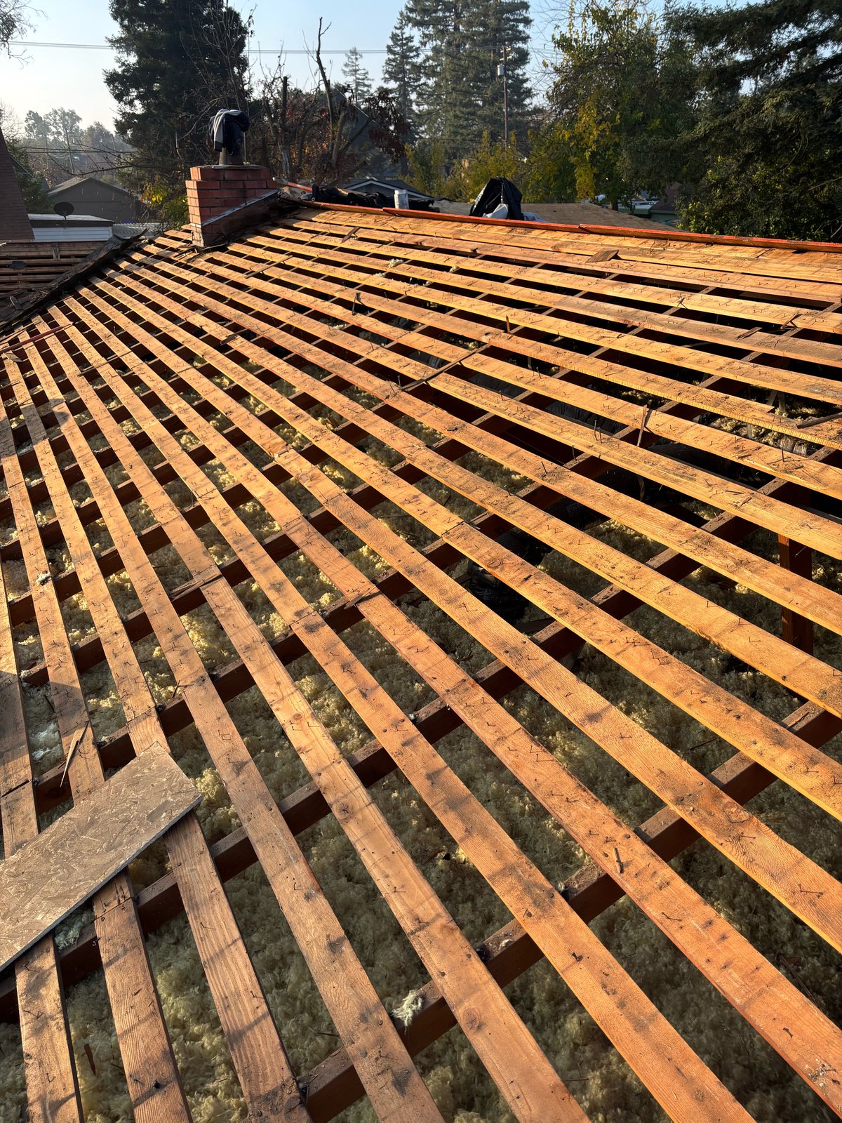 Turlock roof tear-off showing spaced skip sheathing boards over yellow attic insulation beside a brick chimney, with one OSB sheet staged for solid redeck