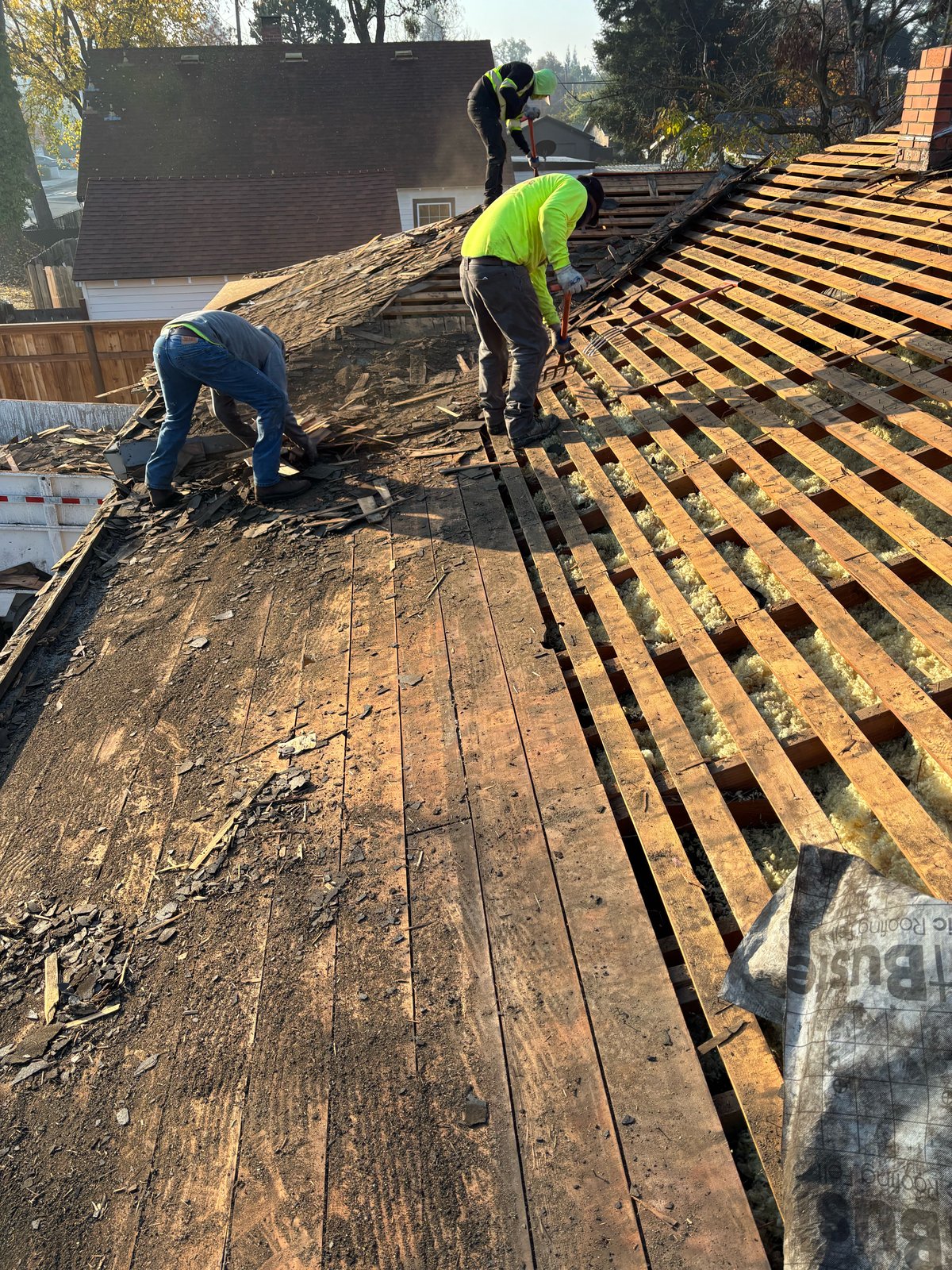 Legend Roofing crew on a Turlock roof tear-off exposing skip sheathing, open rafters, and yellow attic insulation beside a brick chimney