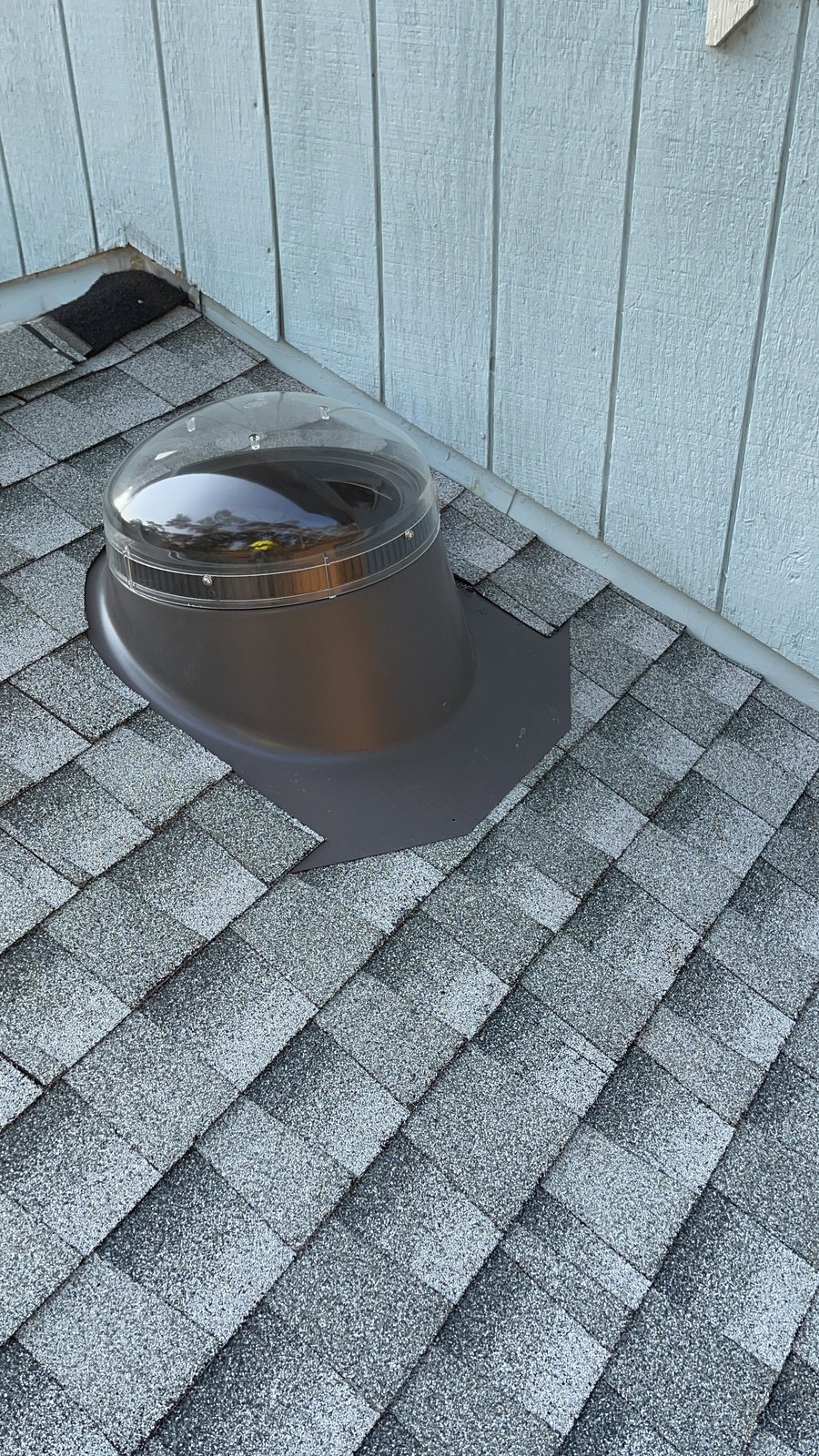 Completed tubular skylight with clear dome and dark metal flashing integrated into charcoal architectural shingles near a light exterior wall