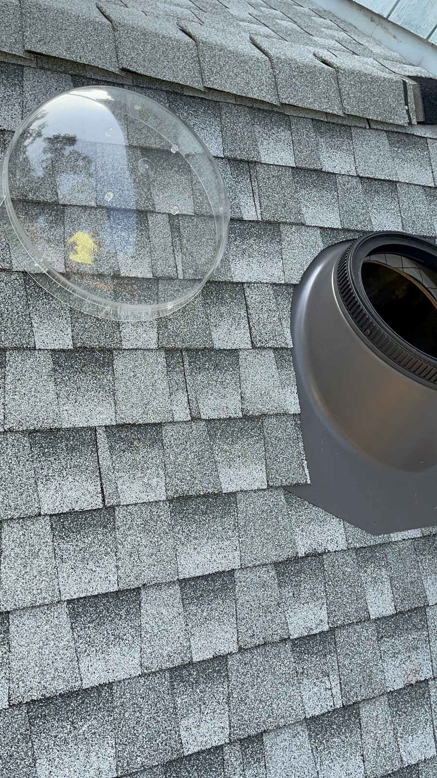 Clear acrylic sun tunnel dome resting on gray shingles beside the installed dark metal flashing base on a Central Valley residential roof