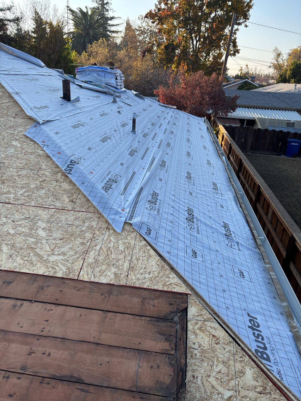 Modesto roof slope with new OSB, GAF FeltBuster synthetic underlayment overlapping toward a metal drip edge at the rake, and two black vent pipes