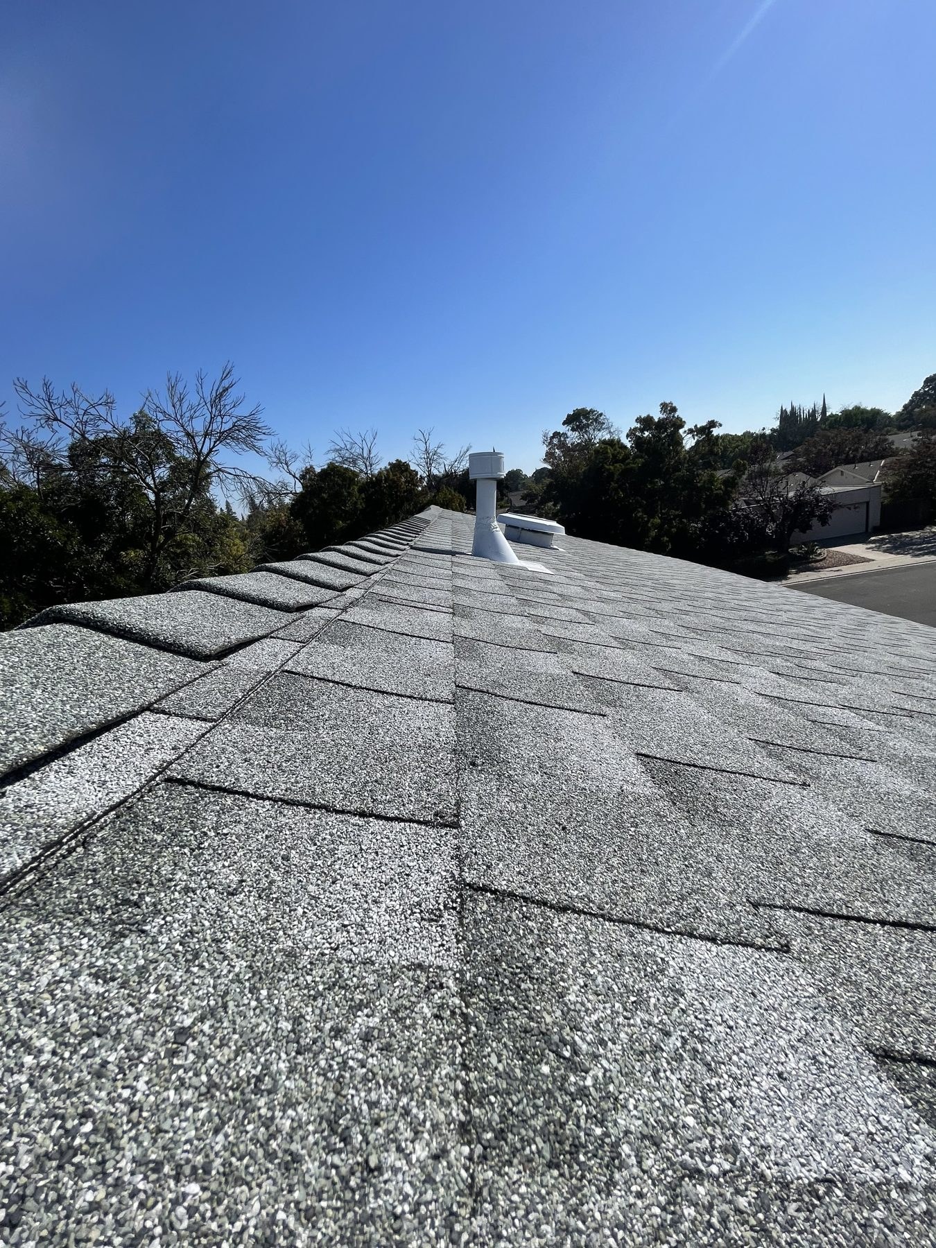 Close-up of gray architectural ridge-cap shingles with a white plumbing stack and a low-profile round attic vent on the slope