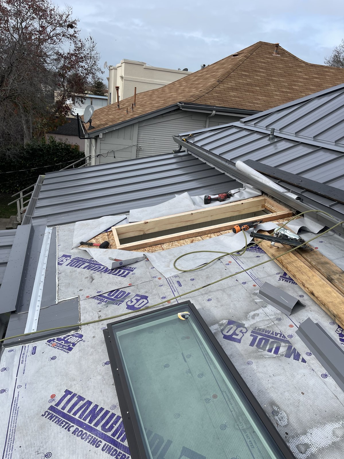 Standing seam metal panels beside Titanium UDL synthetic underlayment with a skylight curb framed open and tools on the roof deck
