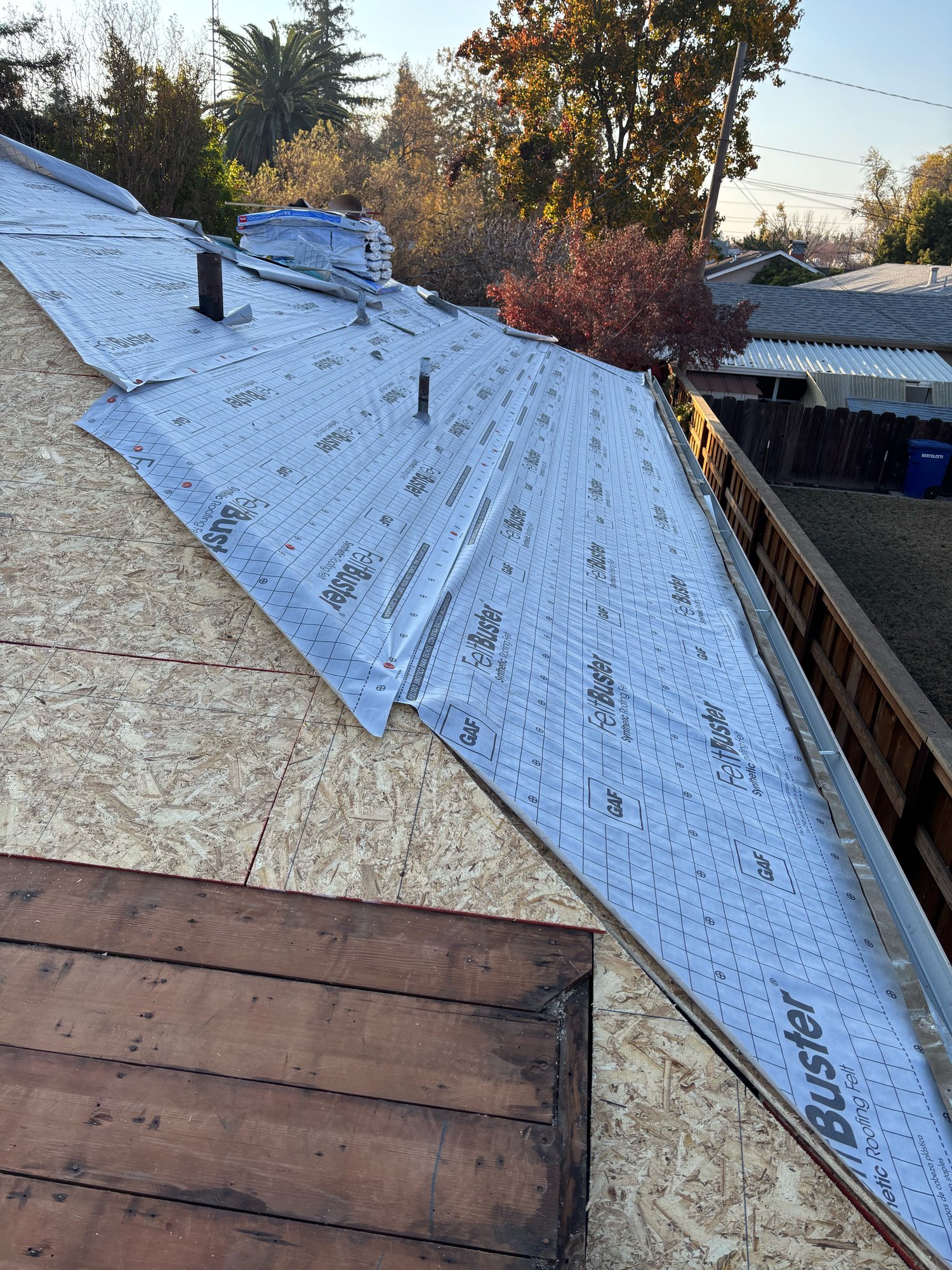 Modesto roof dry-in with GAF FeltBuster synthetic underlayment overlapping toward a shiny galvanized metal drip edge at the rake, new OSB decking, and two black vent pipes