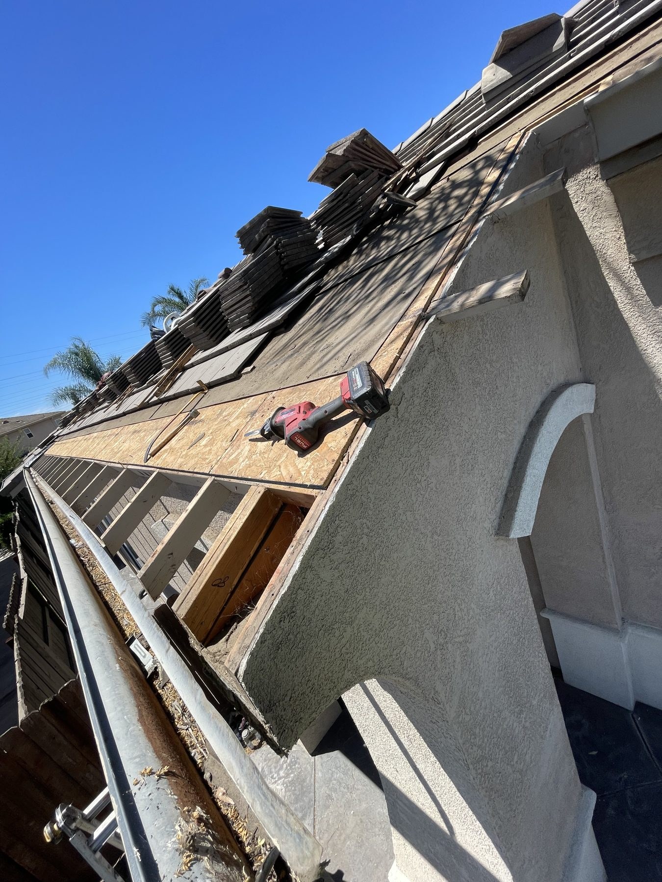 New OSB roof decking installed over exposed rafters with stacked concrete tiles and a Milwaukee reciprocating saw resting near the eave gutter