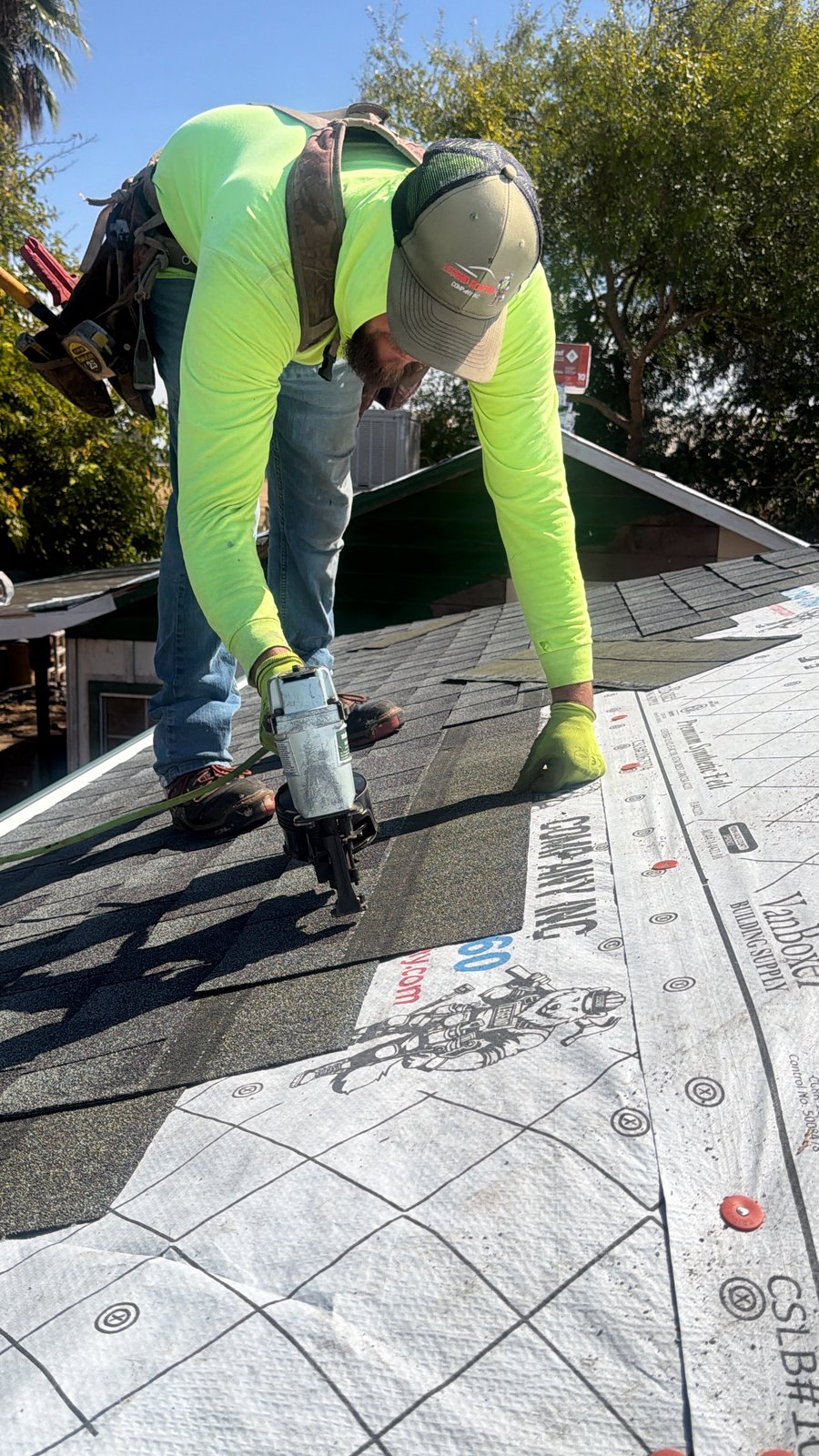 High-angle view of a Legend crew member driving a pneumatic nailer into a dark architectural shingle course over white Legend-branded synthetic underlayment with red plastic-cap nails and printed fastener targets