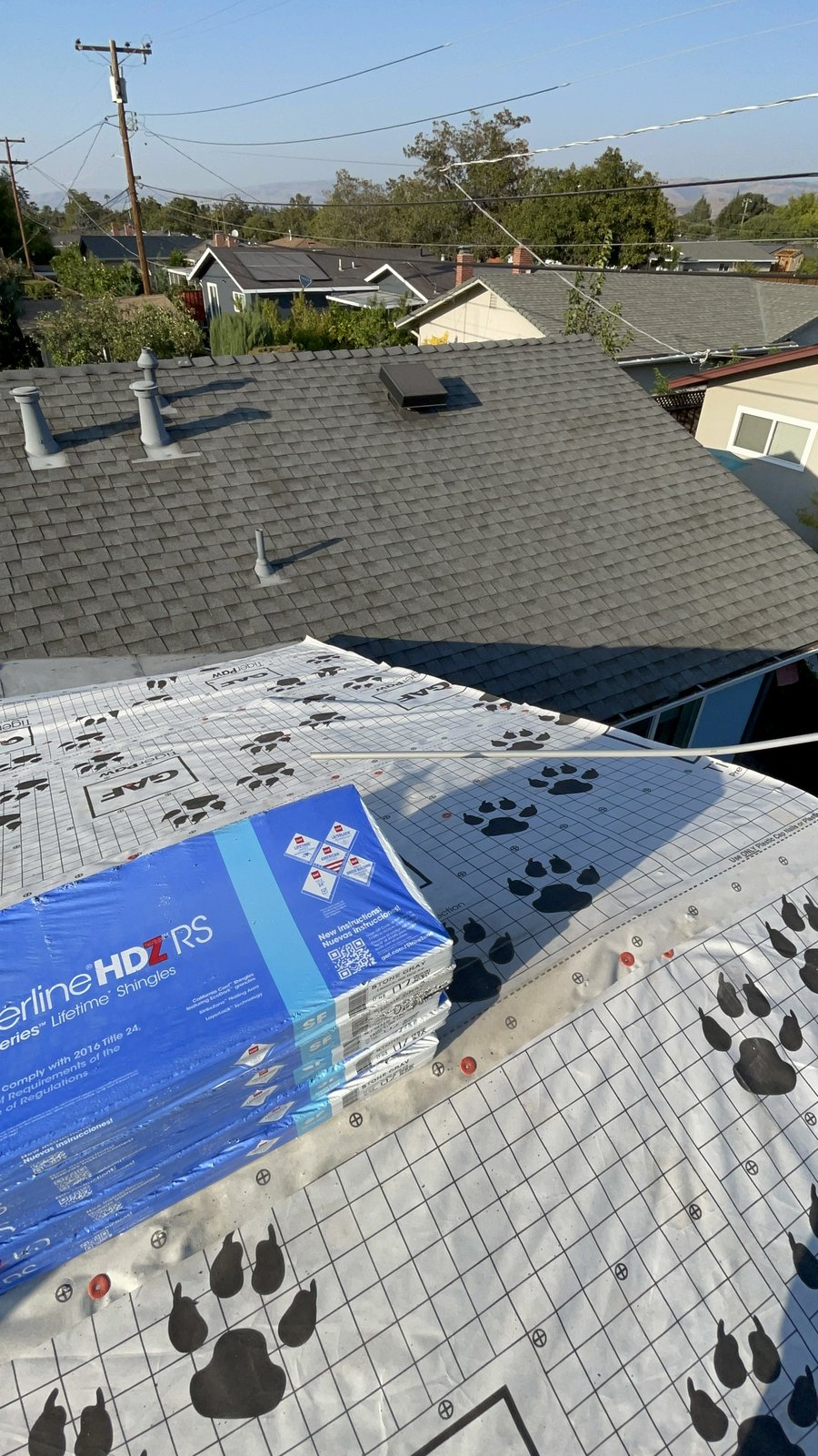 White GAF Tiger Paw synthetic underlayment with red plastic-cap nails and stacked blue Timberline HDZ RS Stone Gray shingle bundles beside a finished gray architectural shingle slope with pipe boots and a box vent
