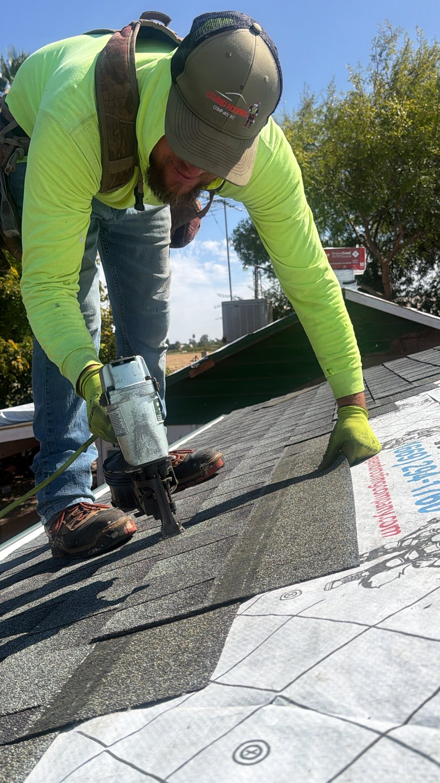 Legend Roofing Company Inc crew member in a branded Legend trucker cap and neon shirt fastening architectural shingles with a coil nailer over underlayment printed with (669) 294-1760 and legendroofingcompany.com