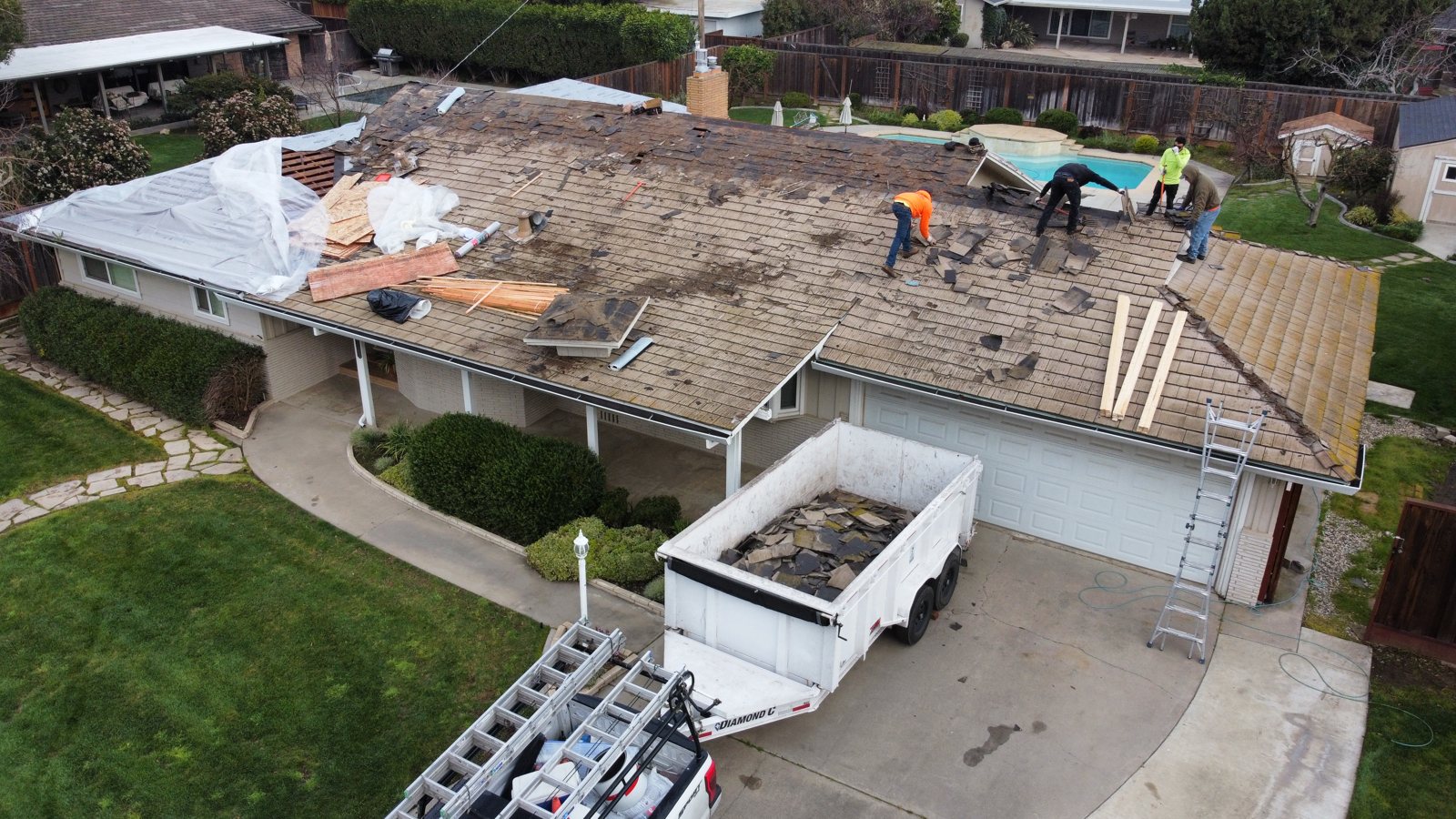 Crew stripping old tile and battens from a Modesto roof into a dump trailer in the driveway, with new sheathing staged on the roof
