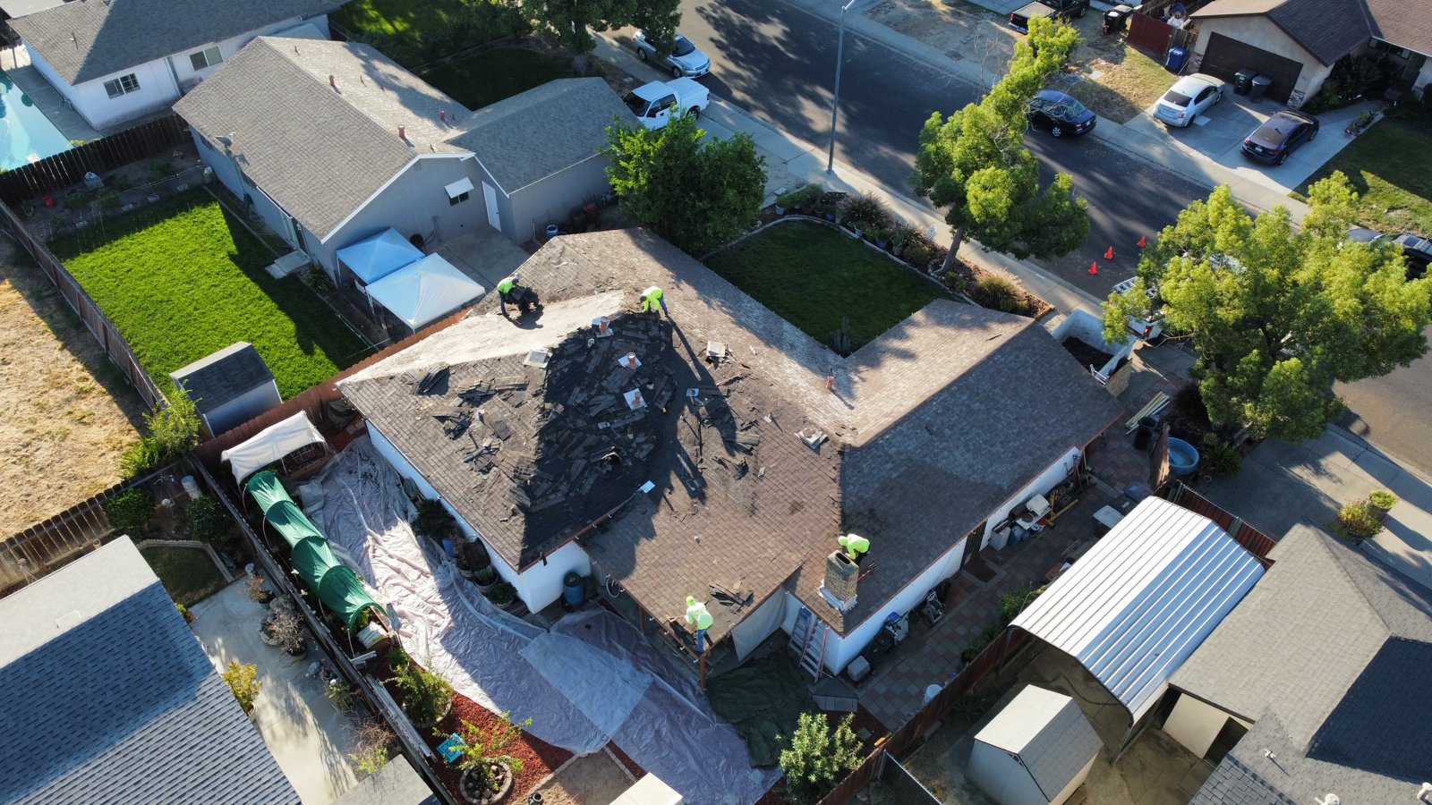 Overhead drone view of the same Riverbank roof with more of the old shingles stripped, exposing the roof deck, as crew members work across several planes