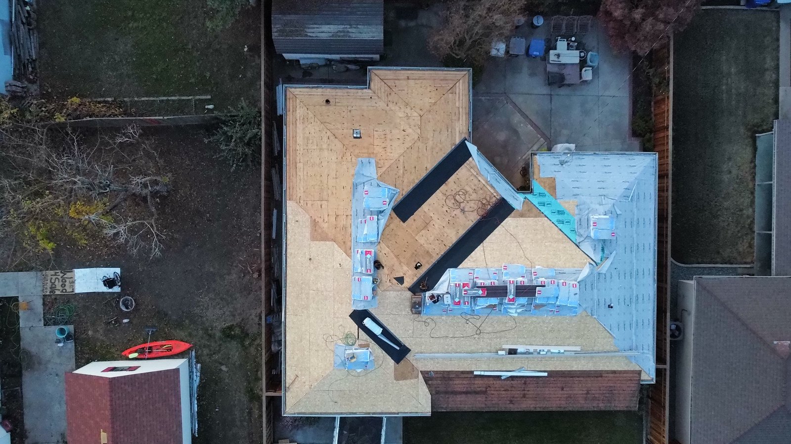Top-down drone view of the same Modesto roof with fresh OSB sheathing, dark membrane in the valleys, synthetic underlayment starting on one side and GAF Grand Sequoia bundles staged along the hips and ridges