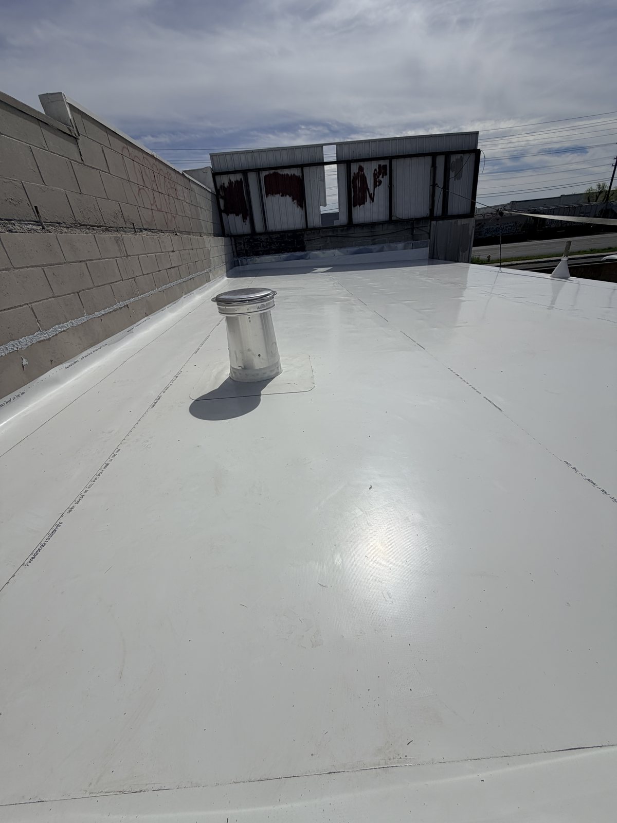 Metal vent stack on a white TPO roof, flashed into the membrane at its base, with welded seams running across the roof toward a parapet wall