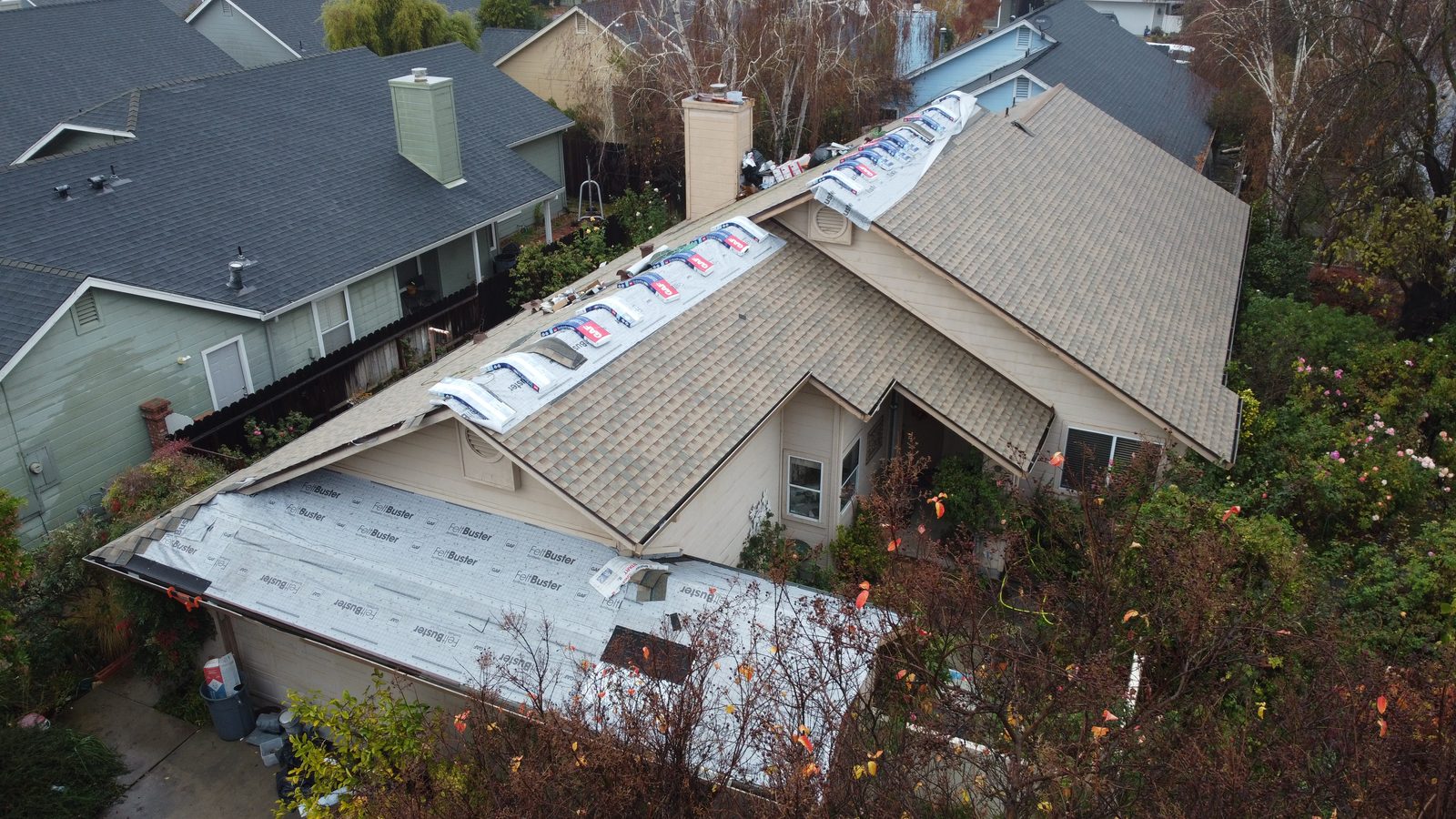 Drone view of a Modesto roof with new light-brown architectural shingles on the main planes, GAF FeltBuster still covering a lower garage section, and shingle bundles staged on the ridge