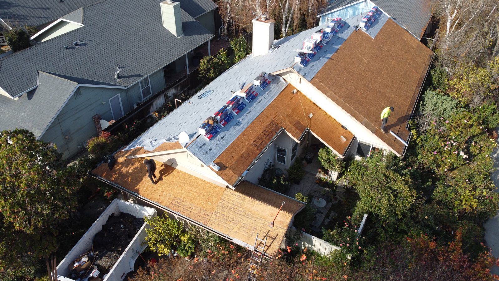Drone view of a Modesto home after roof tear-off with open OSB decking, GAF FeltBuster underlayment on the peaks, staged Timberline shingle bundles, and a debris trailer in the driveway
