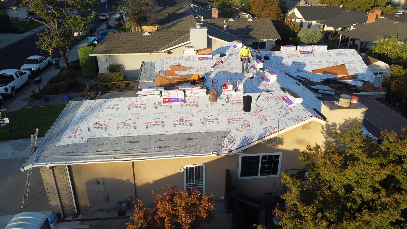 Drone view of a Modesto roof dry-in with Legend Roofing Company Inc branded underlayment, GAF FeltBuster sections, staged GAF shingle bundles, and two crew members on the deck
