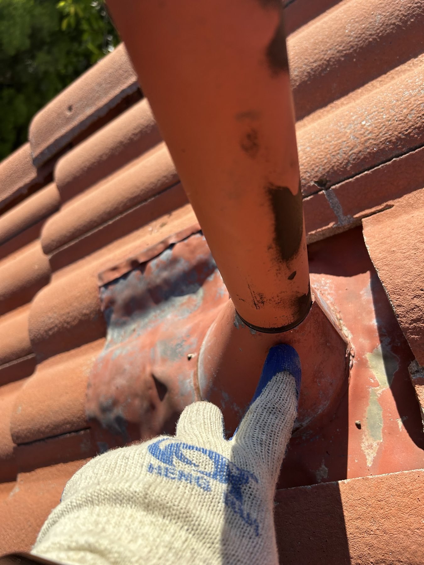 Gloved hand pointing at the base of a painted vent pipe and its worn metal flashing on a clay tile roof