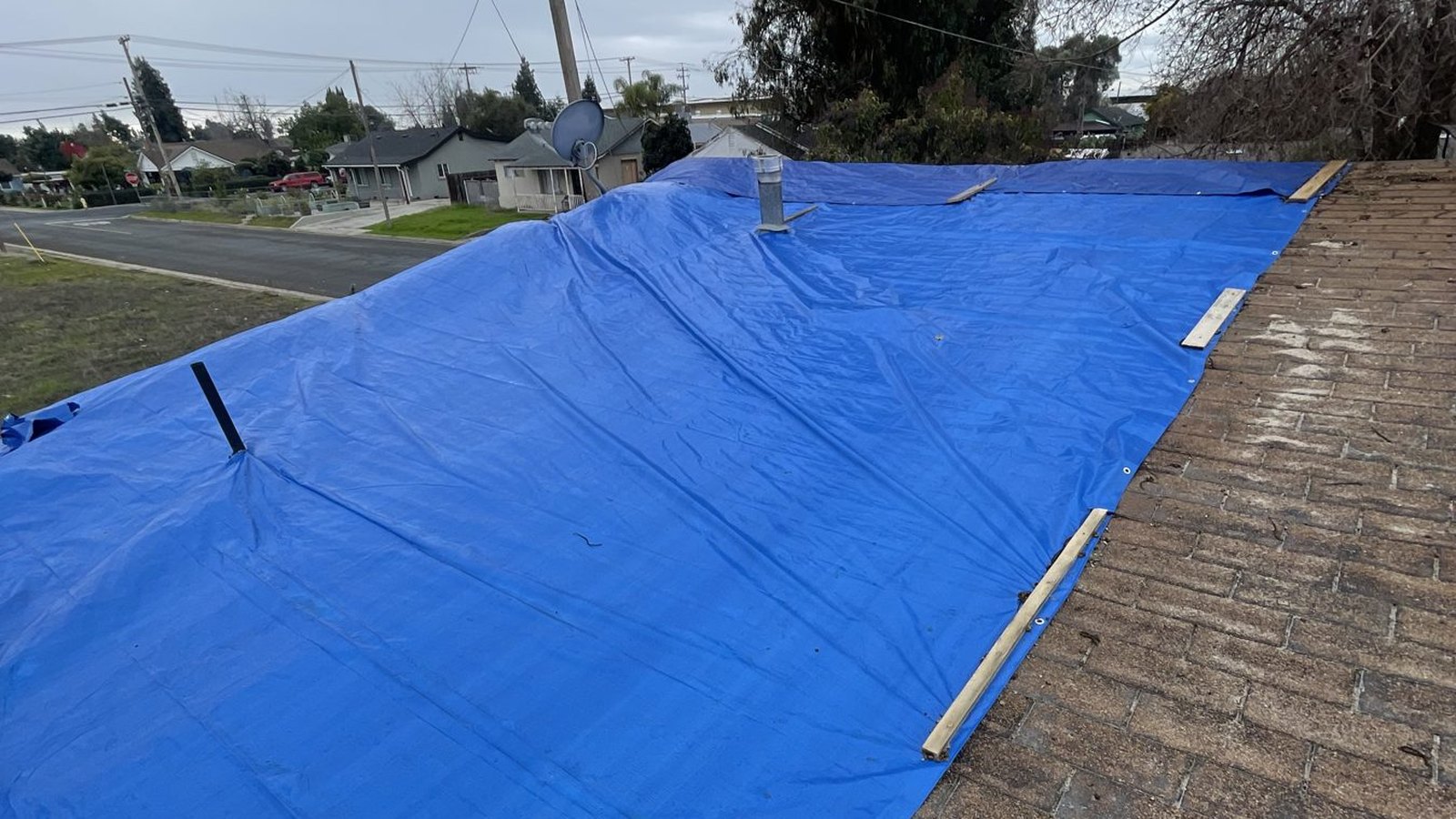 Blue temporary tarp held down with wood battens across a section of an asphalt shingle roof on a residential street under a cloudy sky