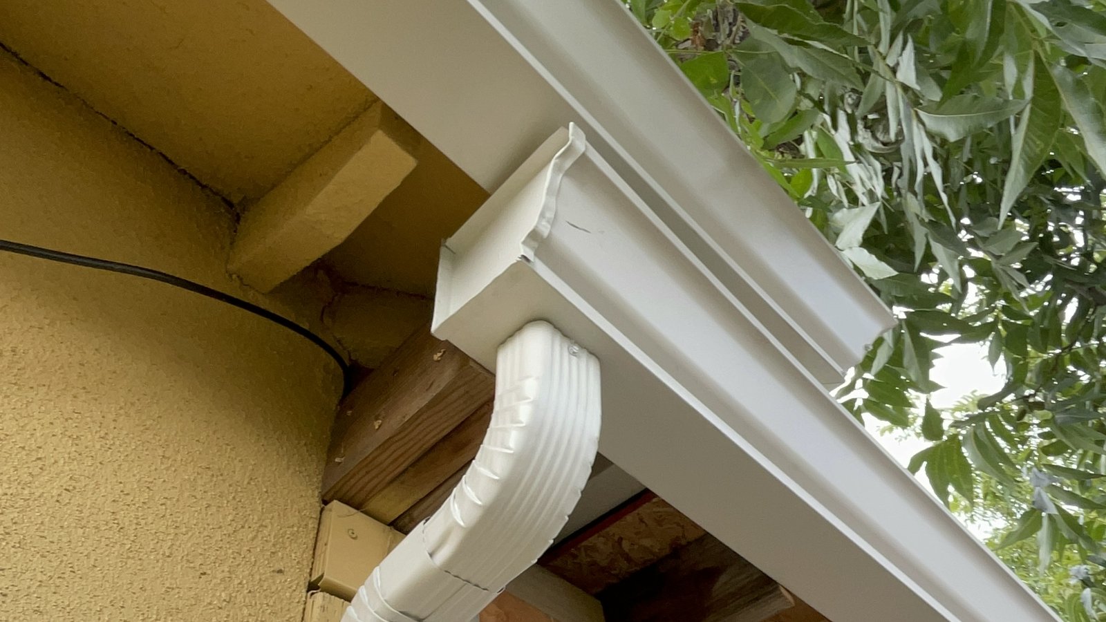 White gutter outlet and downspout elbow under a roof eave on a stucco house