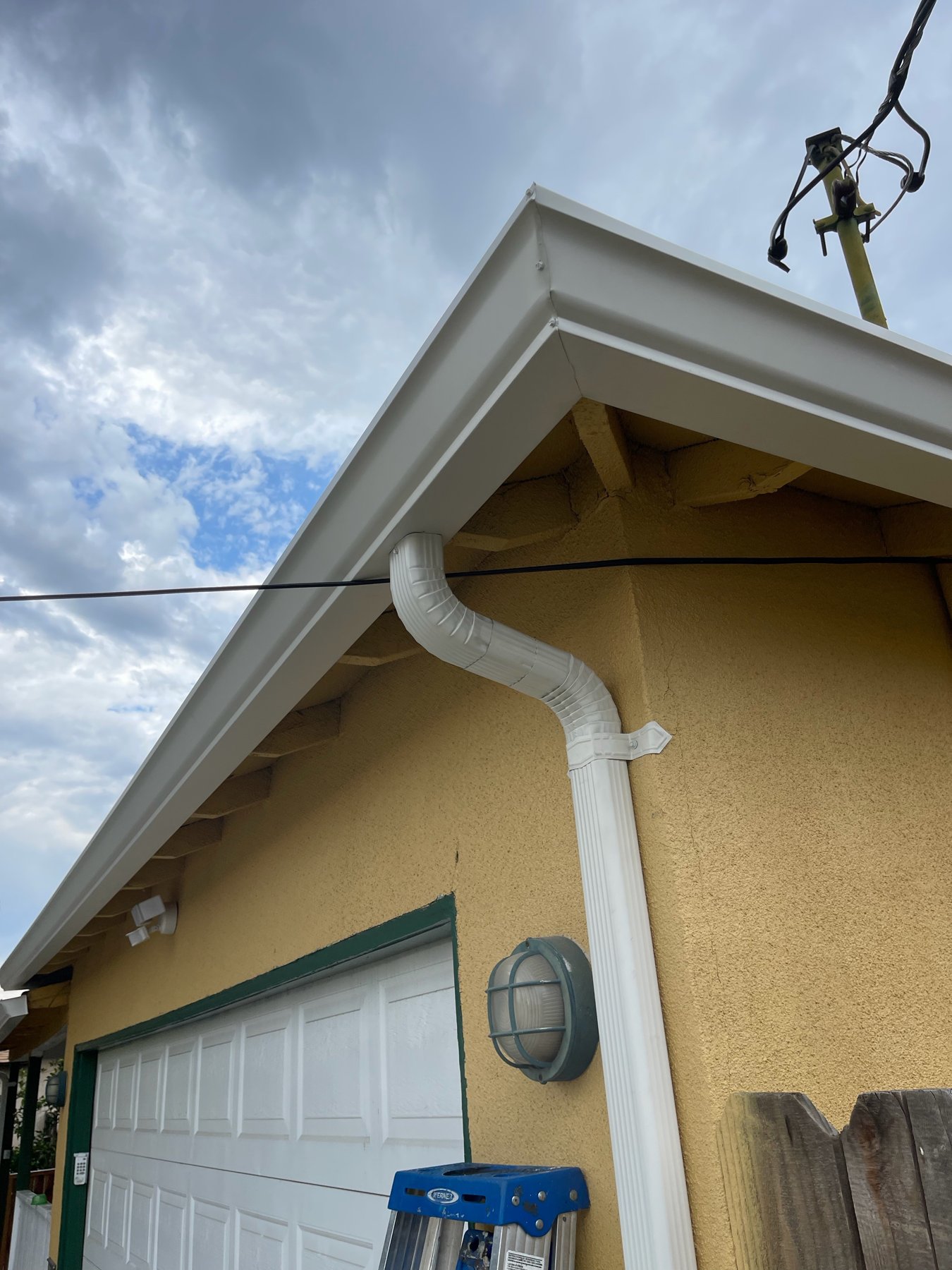 White gutter corner and downspout on a tan stucco garage with a green-trimmed garage door under a cloudy sky