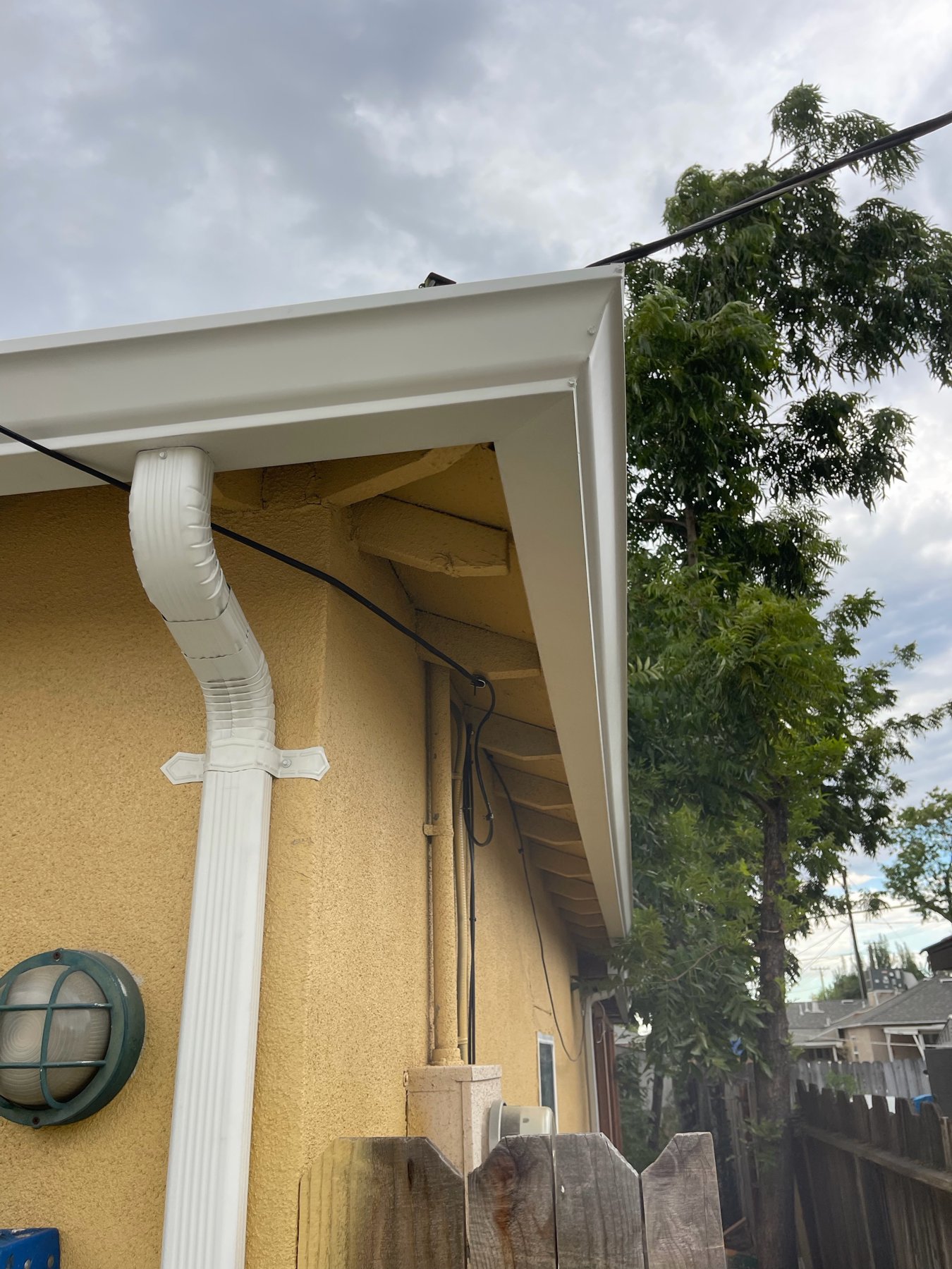 White gutter end and downspout at the corner of a tan stucco house with trees and a wood fence behind it