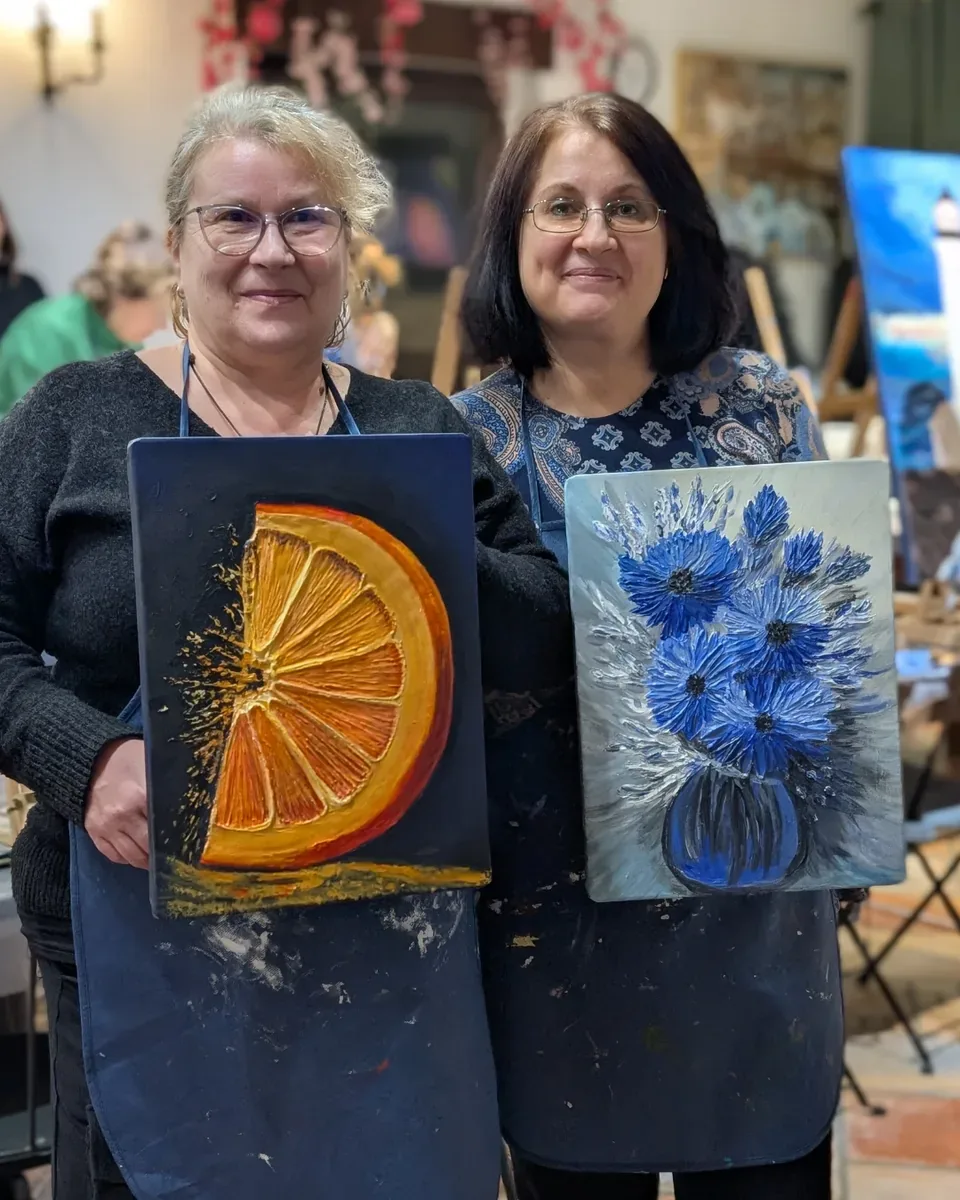 Două cursante zâmbesc în atelier, fiecare cu tabloul ei în relief: o felie de portocală pe fond albastru închis și un buchet de flori albastre într-o vază.