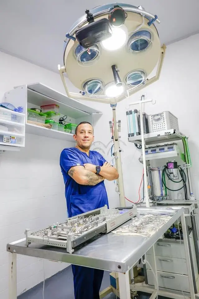 Médico veterinario de uniforme azul, con los brazos cruzados, en el quirófano de Clinica Veterinaria Vital Villeta, bajo la lámpara quirúrgica y junto a la mesa de instrumental.