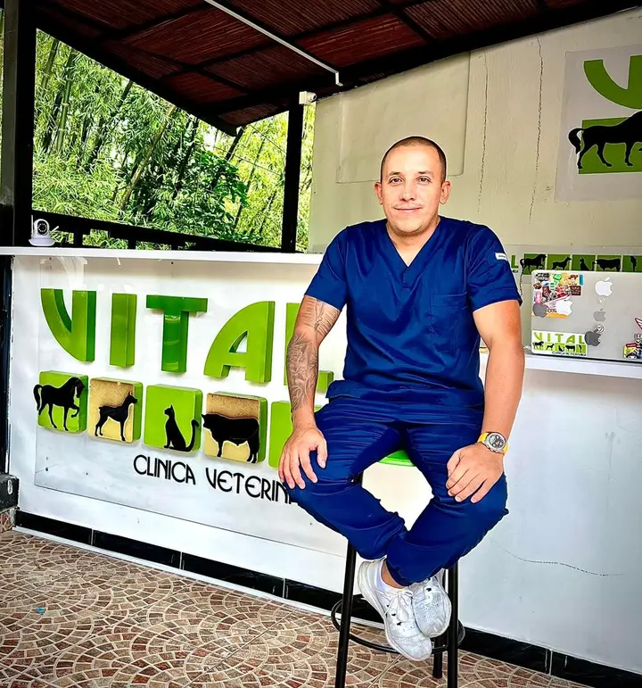 Médico veterinario de uniforme azul sentado frente al mostrador de Clinica Veterinaria Vital Villeta, con el letrero VITAL y la vegetación al fondo.