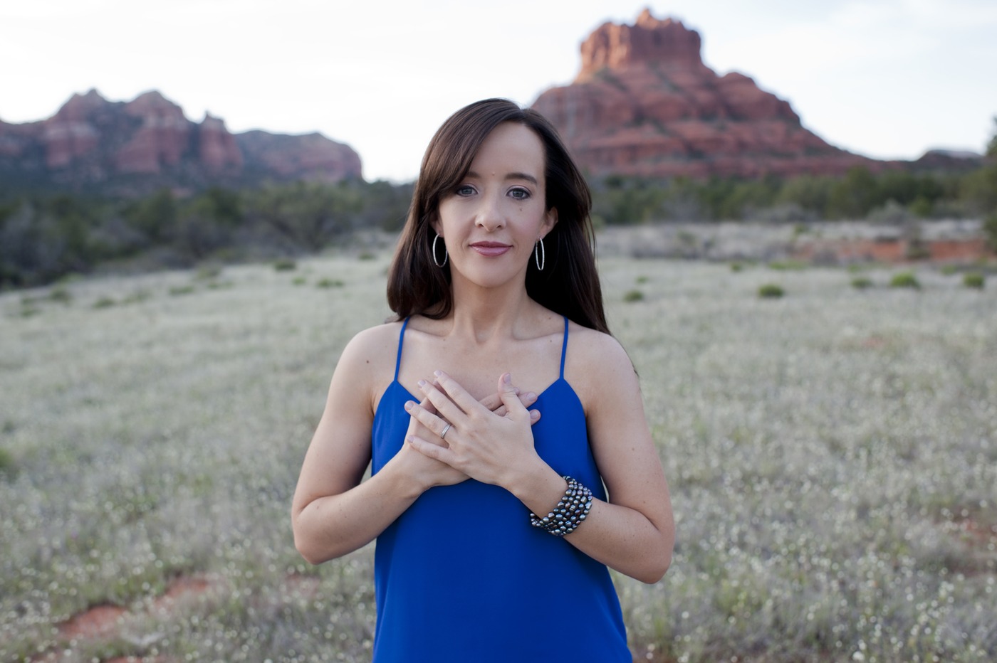 Mindie Kniss in a blue dress with both hands over her heart, standing in a wildflower field with Bell Rock behind her