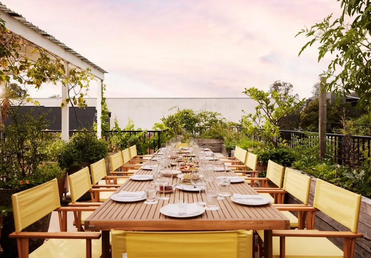 A terrace long table set for service at golden hour, yellow chairs and greenery either side