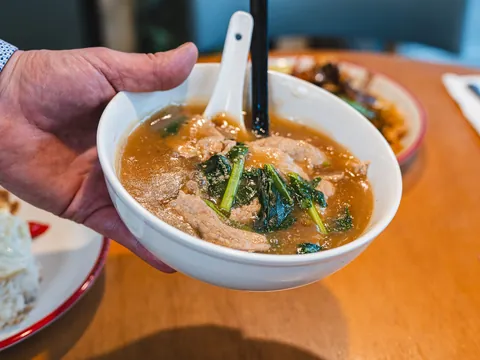 A hand holding up a bowl of noodle soup with greens over a laid table