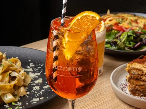 An orange spritz with a straw and an orange slice beside plates of pasta
