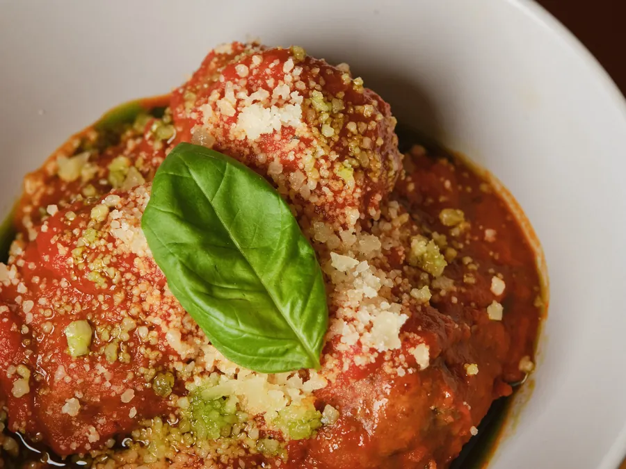 A meatball in tomato sugo under grated cheese, crumb and a basil leaf
