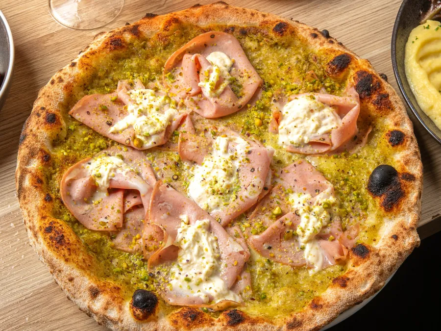 A blistered wood-fired pizza with green sauce, ham and torn burrata on a timber table