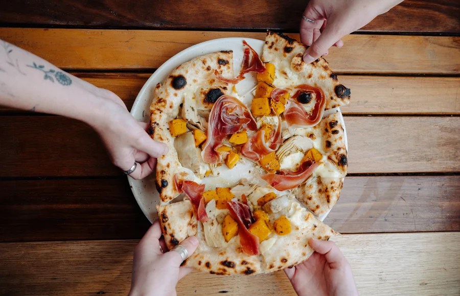 Three pairs of hands pulling apart a pizza on a timber table, shot from above