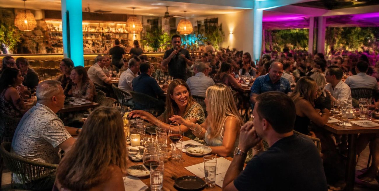A packed restaurant dining room with a host on the mic and teal and violet uplighting